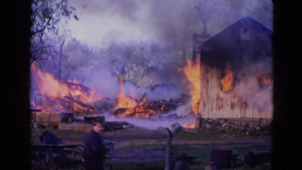 1968 Wauconda, Illinois frame with a person watching from the foreground as flames burn debris and a wooden structure beyond a fence.