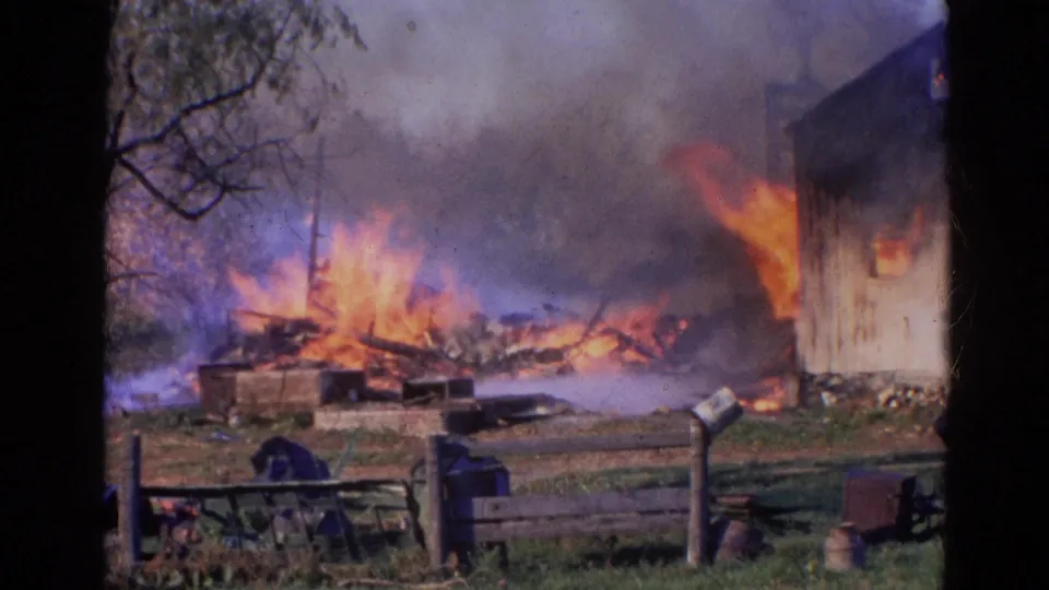 1968 Wauconda, Illinois 8mm frame showing heavy flames and smoke across debris, with a wooden building edge and fence in view.