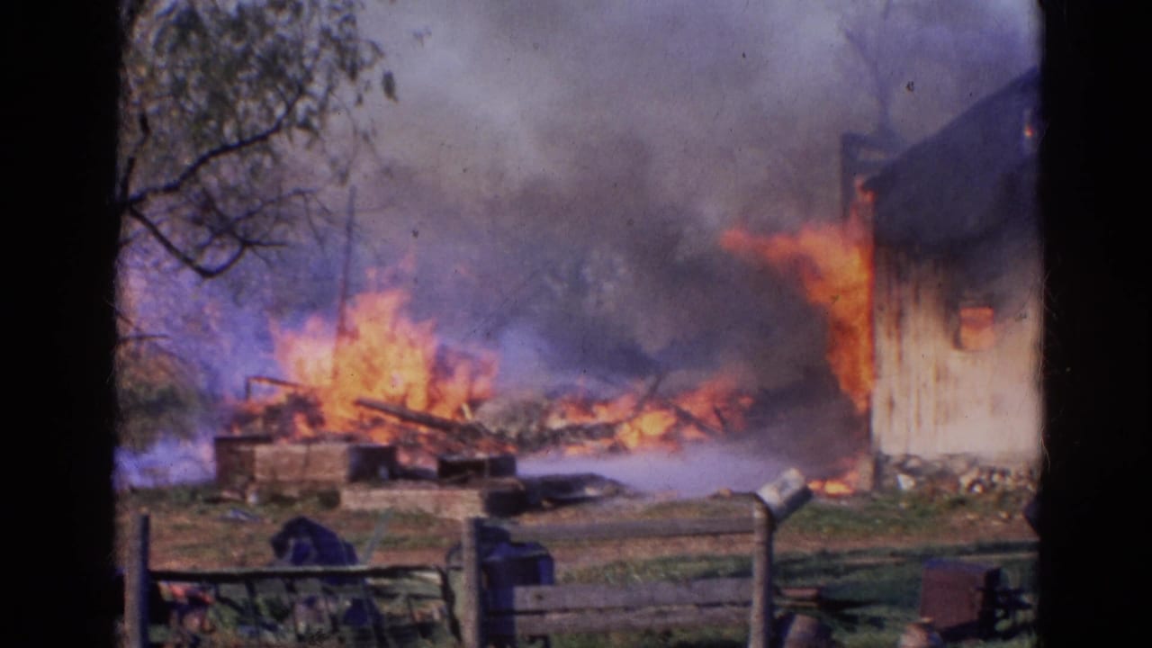 Flames and smoke engulf a rural wooden structure behind a fence.