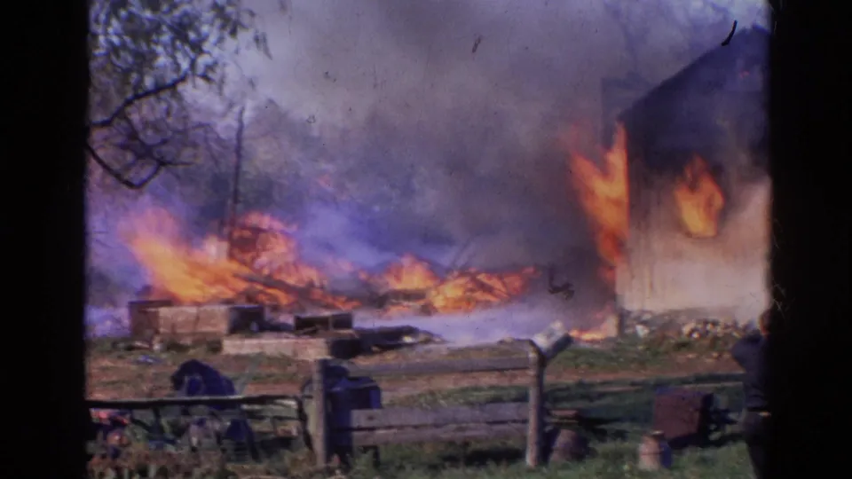 1968 Wauconda, Illinois frame with fire along a wooden building wall, burning debris, smoke, trees, and yard fencing.