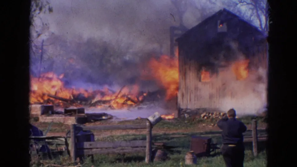 1968 Wauconda, Illinois 8mm frame showing a dark-clothed observer near a fence as a rural building and debris burn.