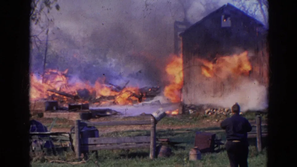 1968 Wauconda, Illinois frame showing a bystander facing a structure fire with heavy smoke, flames, fencing, and scattered yard items.