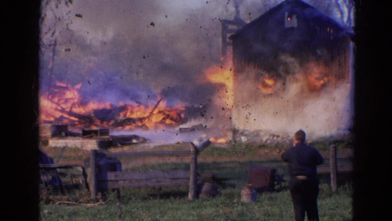 Large flames and smoke rise from a rural structure with a person watching from the right.