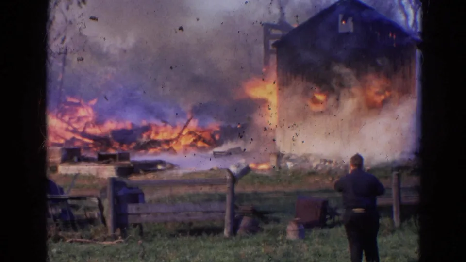 The barn fire continues in Wauconda, Illinois, with flames along the structure, smoke overhead, fencing, and a lone figure in the foreground.