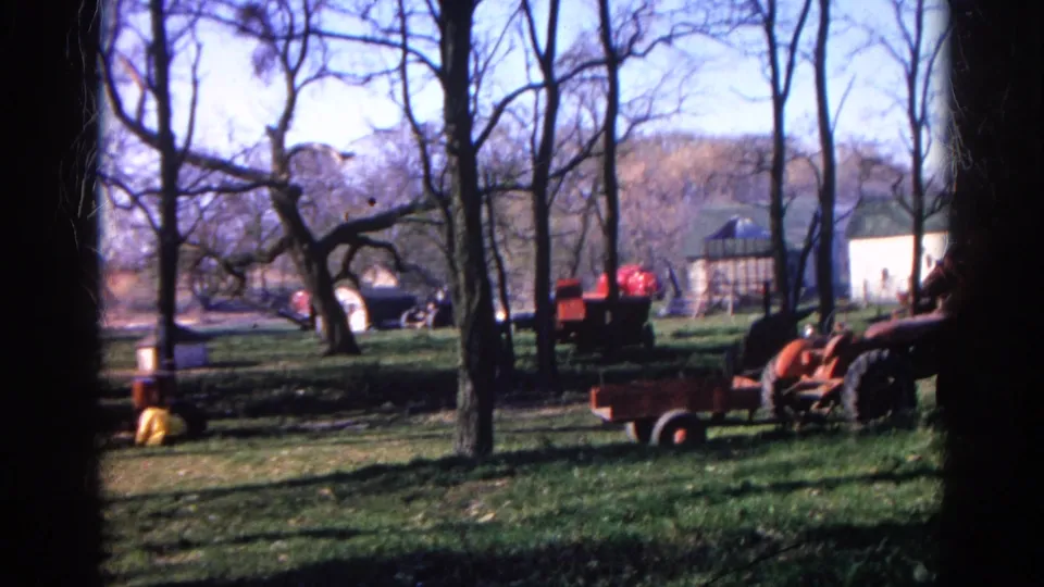 A rural Wauconda yard with leafless trees, red tractor equipment, wagons, and small outbuildings in faded 1968 8mm color.