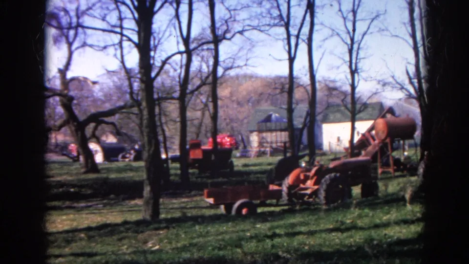 Old red farm equipment, wagons, bare trees, and light-colored outbuildings appear in a rural Wauconda yard on 8mm film.