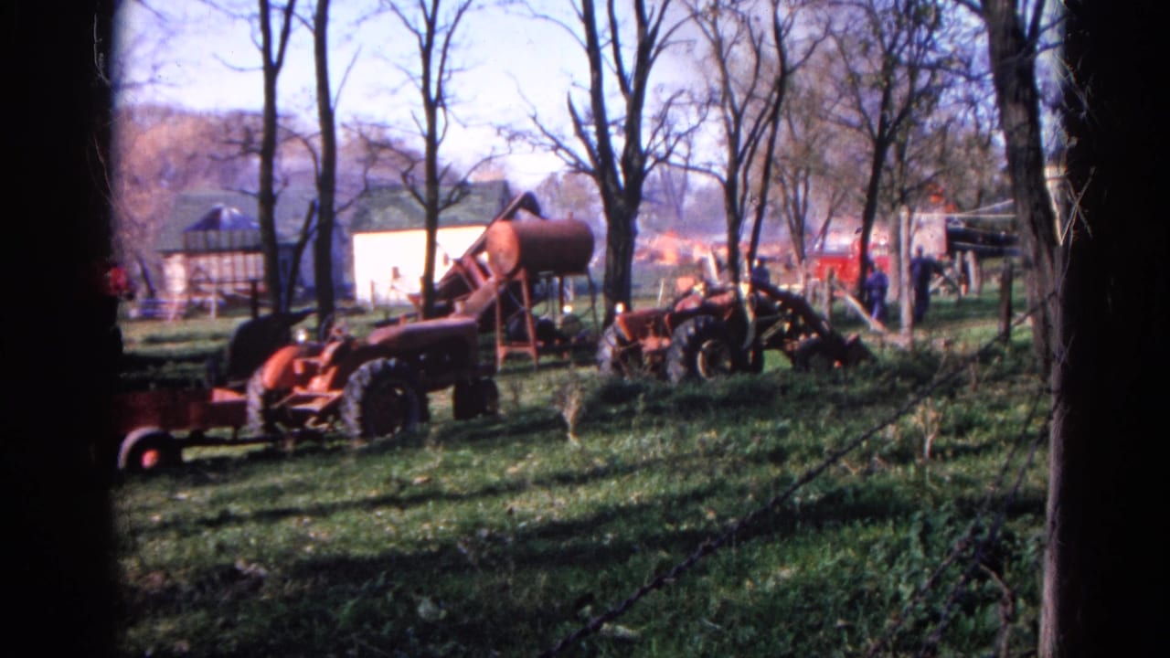 1968 Wauconda farm scene with red tractors, a cylindrical tank, bare trees, and flames near outbuildings in the distance.
