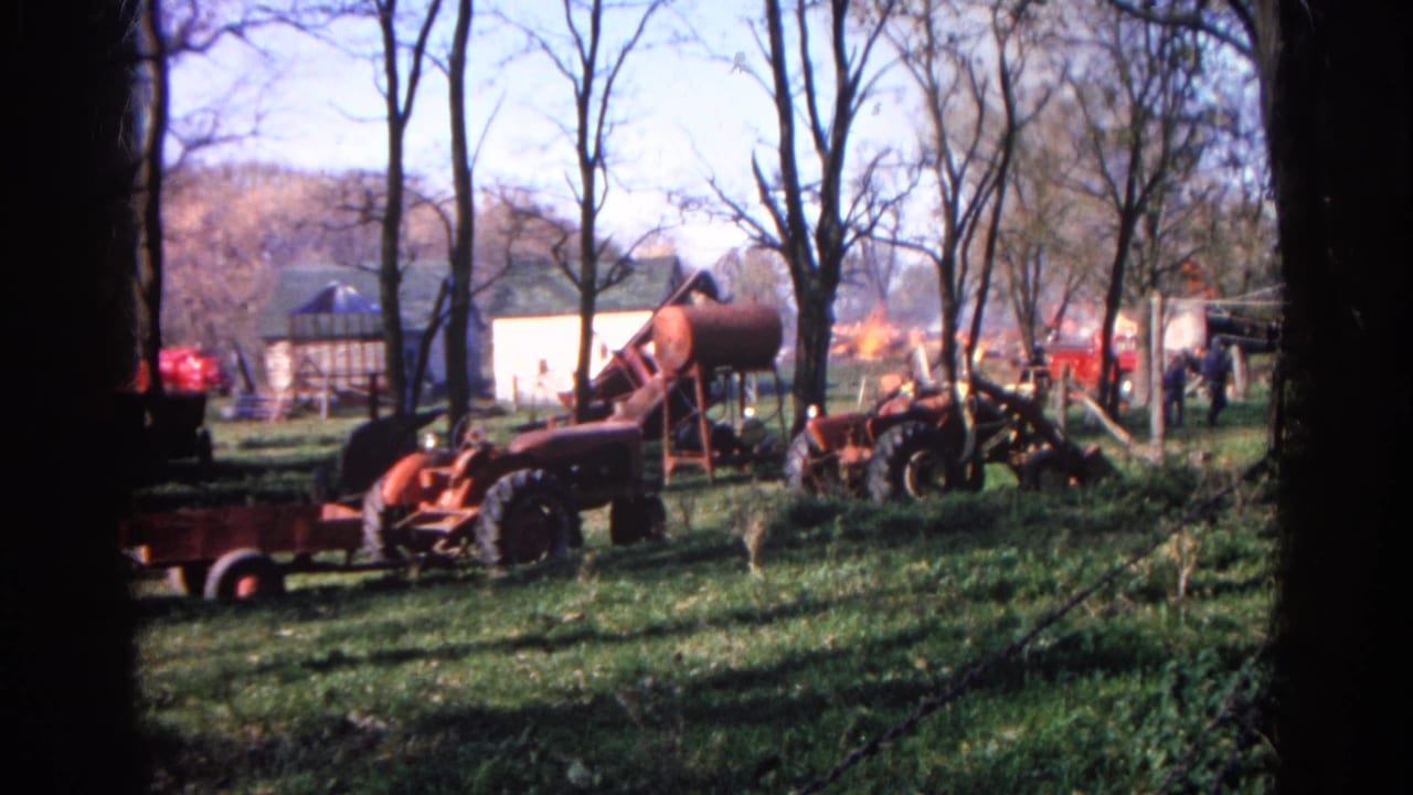Red tractor, tank, trees, and distant onlookers appear on a Wauconda farm as smoke or flame shows behind the buildings.