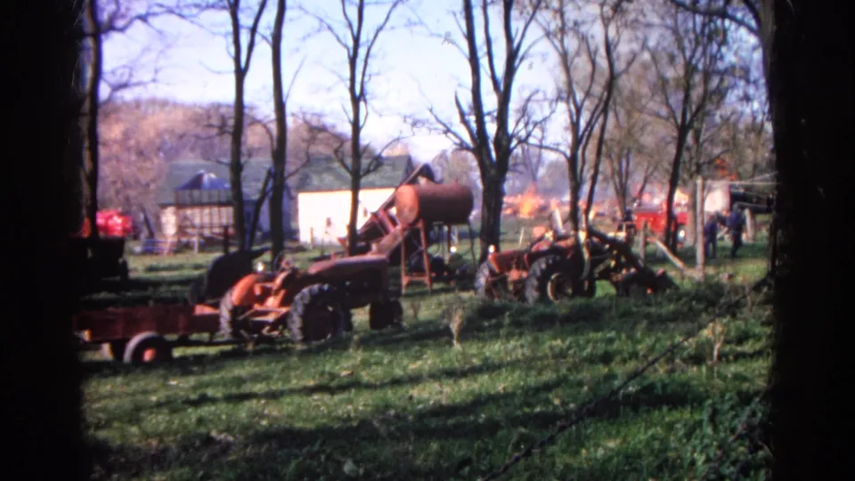 Red tractor, tank, trees, and distant onlookers appear on a Wauconda farm as smoke or flame shows behind the buildings.
