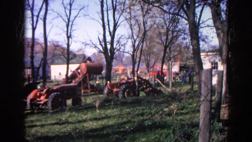 Rural Illinois farm view with red machinery, bare trees, white buildings, and a fire visible in the background.