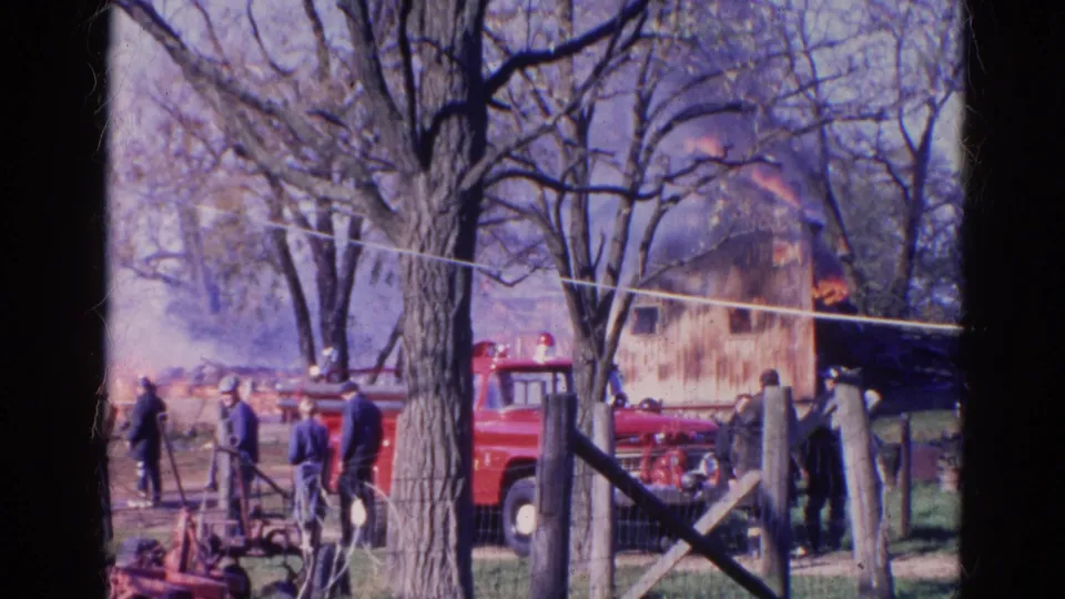 1968 Wauconda, Illinois frame with townspeople, a red fire truck, bare trees, smoke, flames, and a wooden structure in the background.