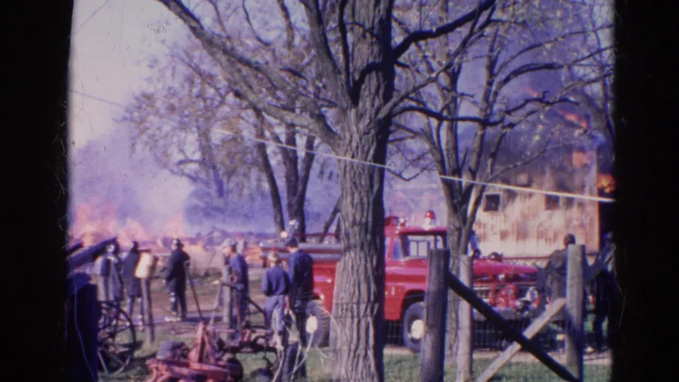 1968 Wauconda, Illinois home-movie frame showing onlookers, a red fire truck, farm equipment, trees, smoke, and flames near a rural structure.