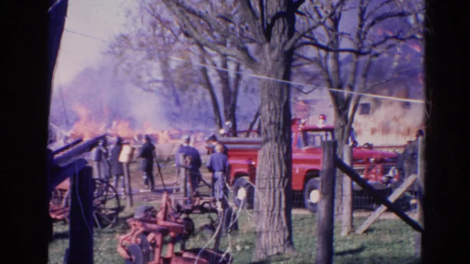 1968 Wauconda, Illinois frame showing a rural fire scene with smoke, flames, a red fire truck, wooden fencing, bare trees, and people watching.