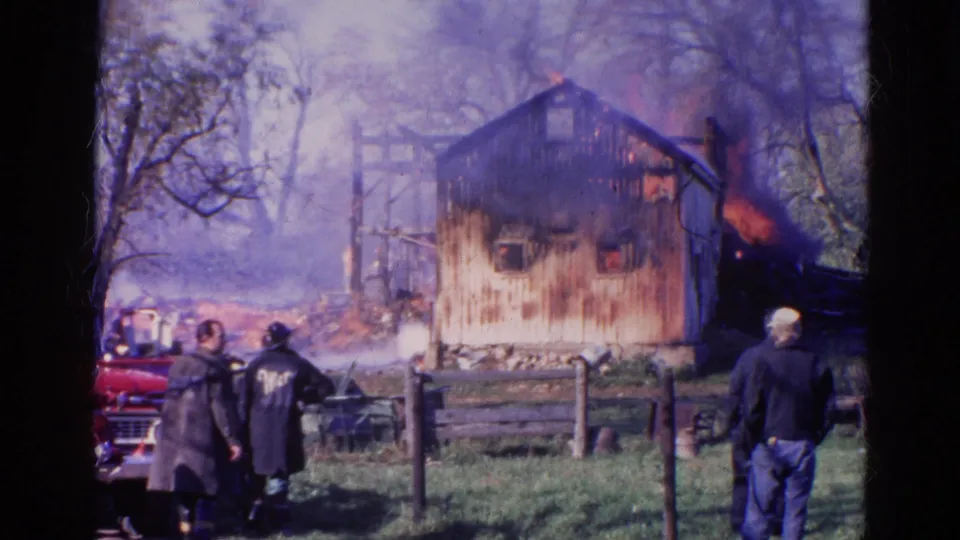 1968 Wauconda home-movie frame showing firefighters and onlookers before a smoking, charred barn wall.
