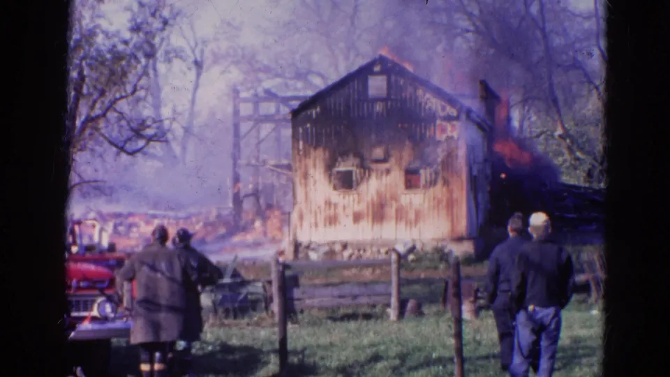 1968 Wauconda frame of a smoking barn fire with people watching from behind a wooden fence.
