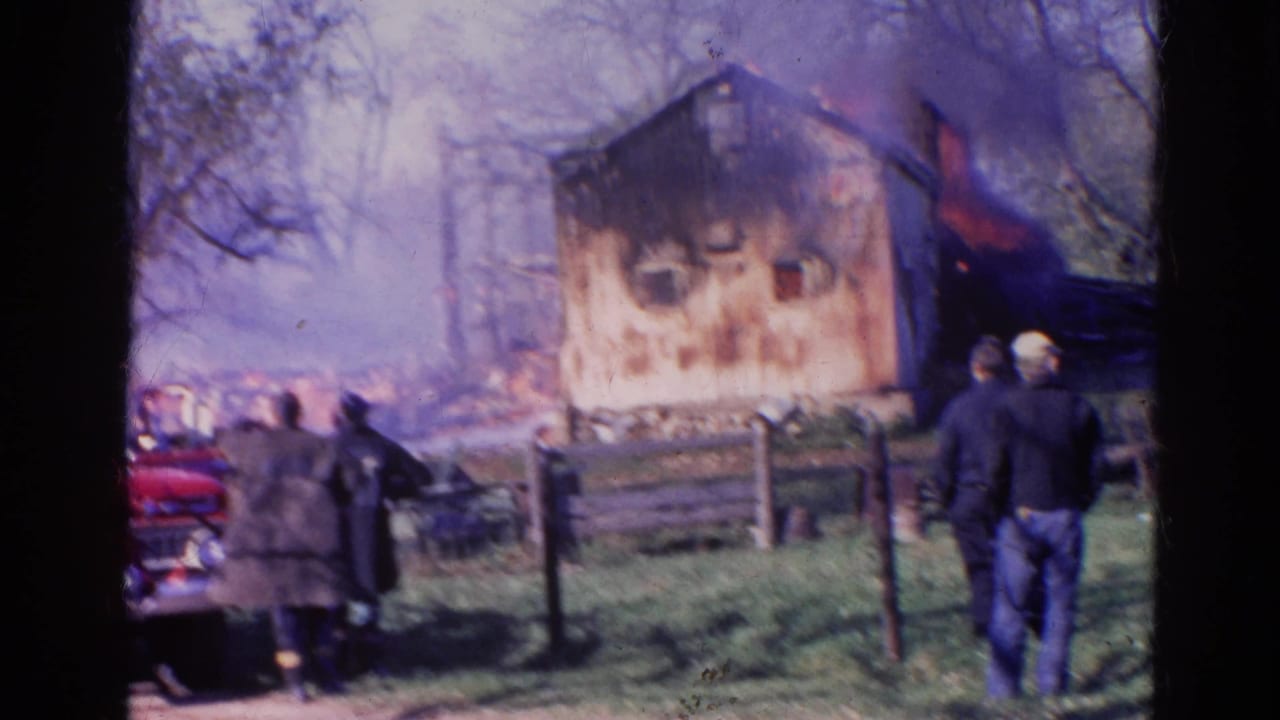 Wooden building burning behind a fence as onlookers stand in a grassy area.