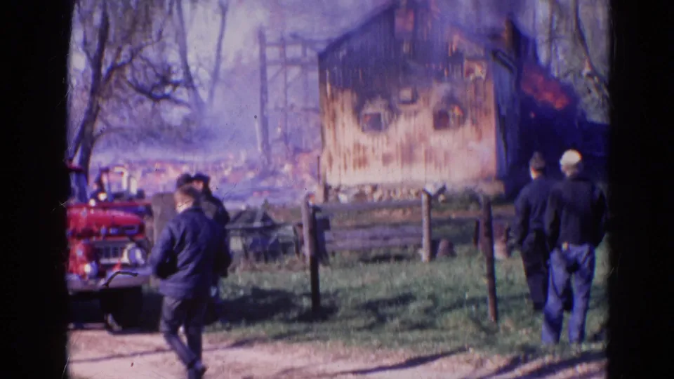 Wauconda 1968 home-movie frame with a red fire truck, responders or onlookers, smoke, fence, and a burning wooden structure.
