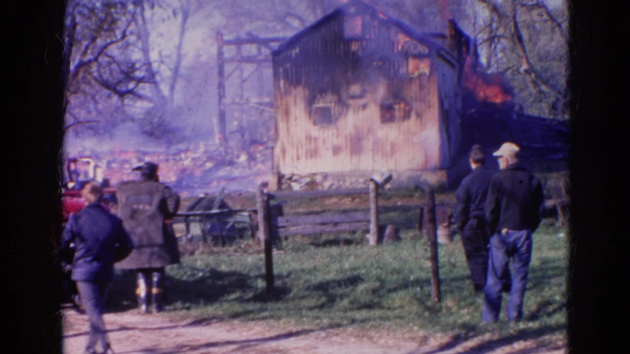 1968 Wauconda, Illinois 8mm frame of adults behind a fence watching a rural wooden structure burn amid smoke and bare trees.