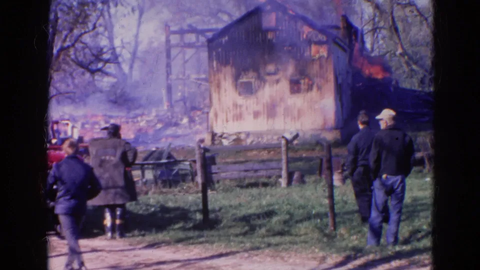 1968 Wauconda, Illinois 8mm frame of adults behind a fence watching a rural wooden structure burn amid smoke and bare trees.