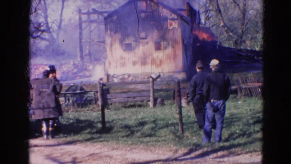1968 Wauconda, Illinois home-movie frame of adult bystanders near a fence looking toward flames on a rural wooden building.