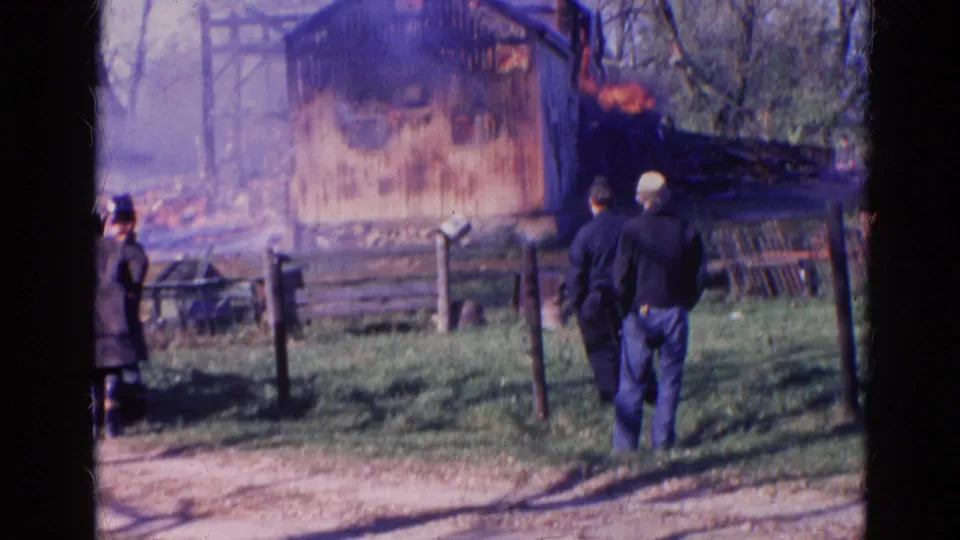 1968 Wauconda, Illinois 8mm frame with two adults in the foreground watching a smoke-filled structure fire behind fencing.