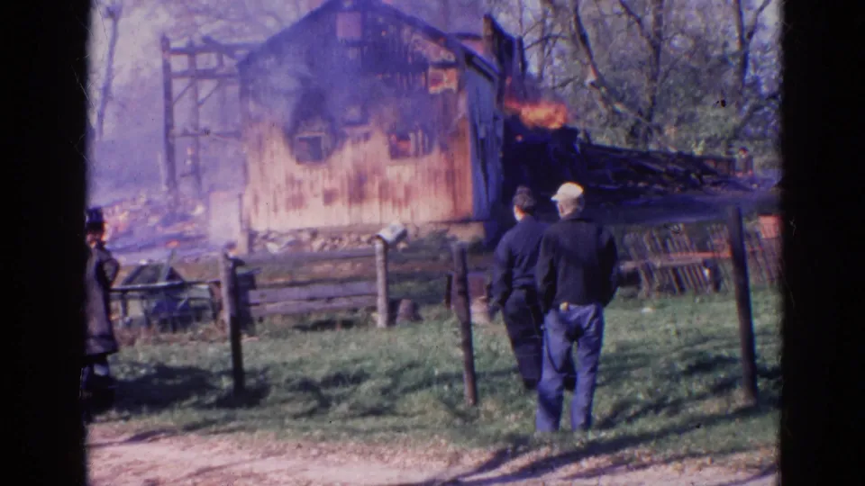 1968 Wauconda, Illinois home-movie frame of two adults watching flames and smoke around a rural wooden structure.