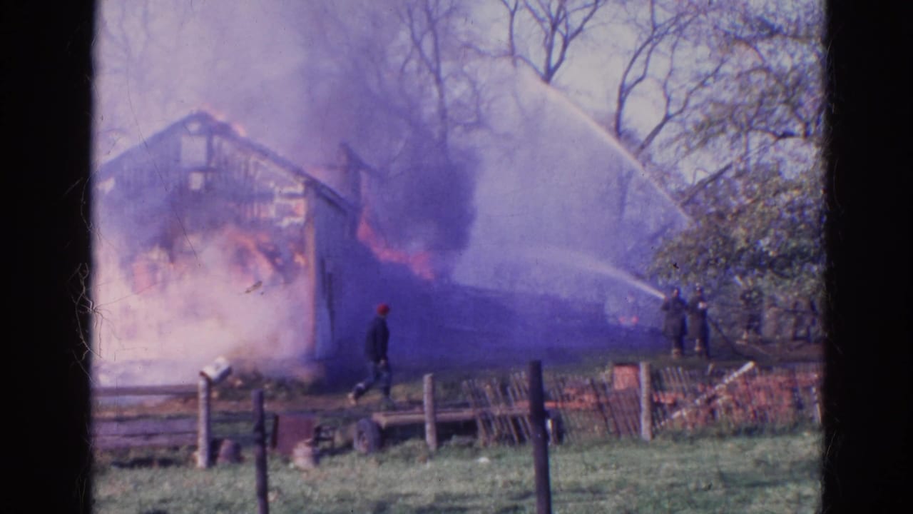 Wooden structure burning as smoke rises and firefighters spray hoses from a grassy field.