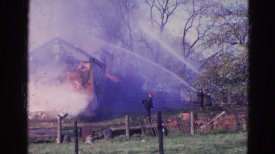 1968 Wauconda, Illinois frame with firefighters in dark clothing aiming hose streams at a burning rural wooden structure.