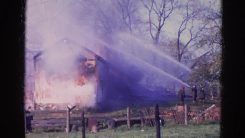 1968 Wauconda, Illinois home-movie frame showing heavy smoke, visible flames, hose spray, firefighters, fencing, and bare trees.