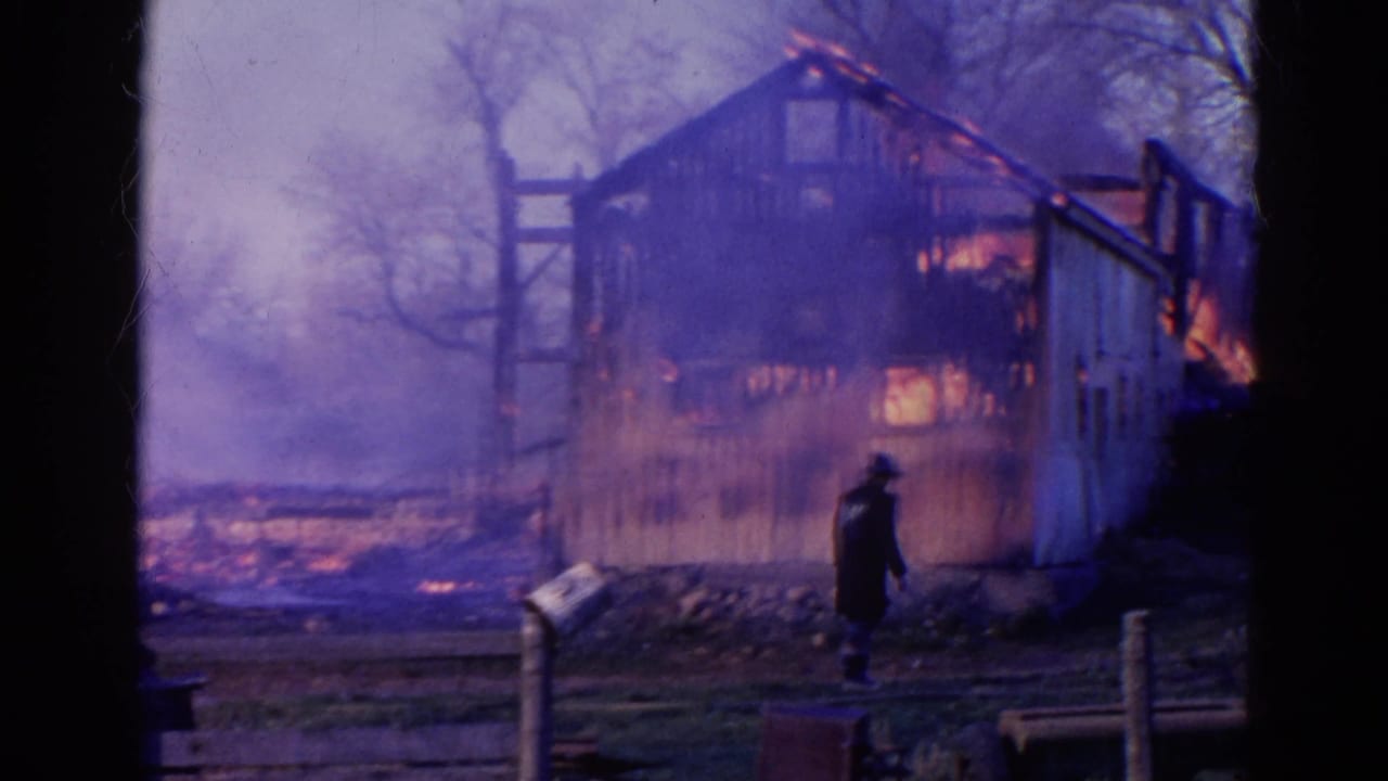 A barn burns with smoke rising as a dark-clothed person walks nearby.