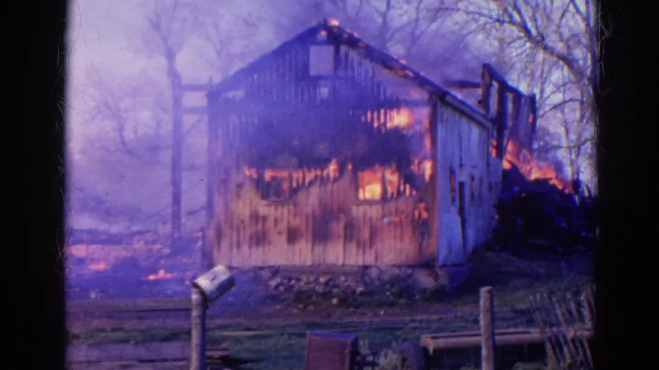 1968 Wauconda, Illinois 8mm frame showing the side of a burning barn, smoke haze, bare trees, and a tilted fence in the foreground.