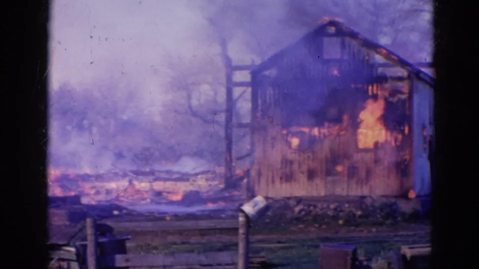 1968 Wauconda, Illinois frame showing a smoke-filled barn fire, bare trees, low flames in the yard, and posts in front.