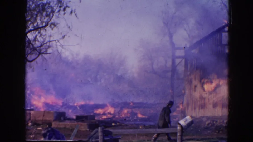 Heavy smoke drifts across the Wauconda fire scene as a dark-clothed figure stands near debris beside the burning structure.