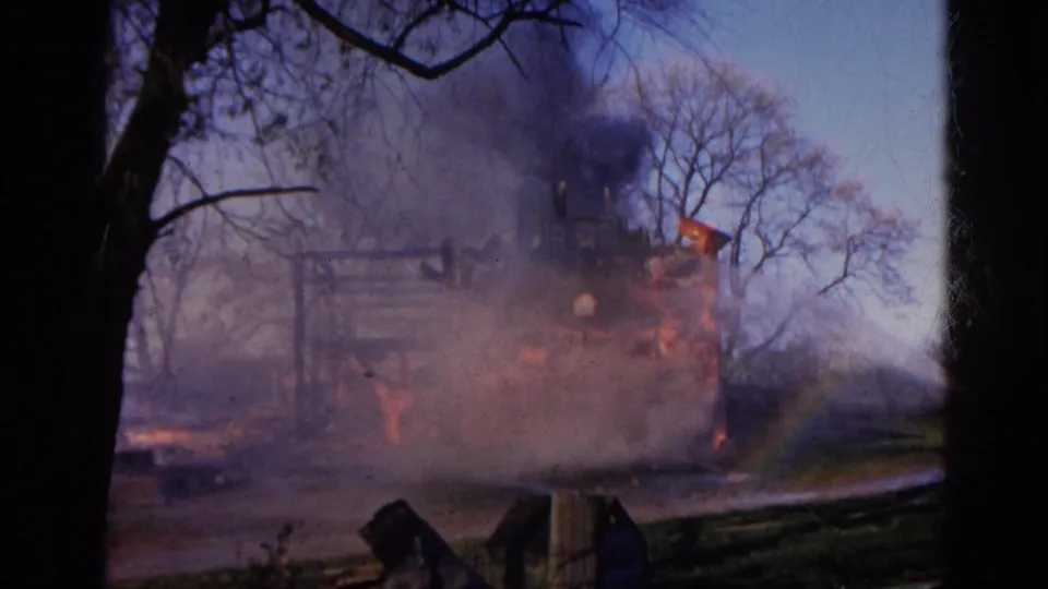 1968 Wauconda 8mm frame of a structure fire with heavy smoke, visible flames, bare trees, and a dark tree trunk in the foreground.