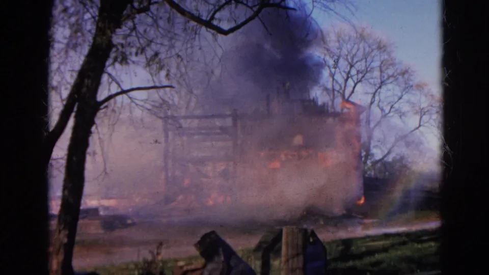 1968 Wauconda 8mm frame showing a smoke-covered building fire, leafless trees, foreground debris, and orange flame along the lower structure.