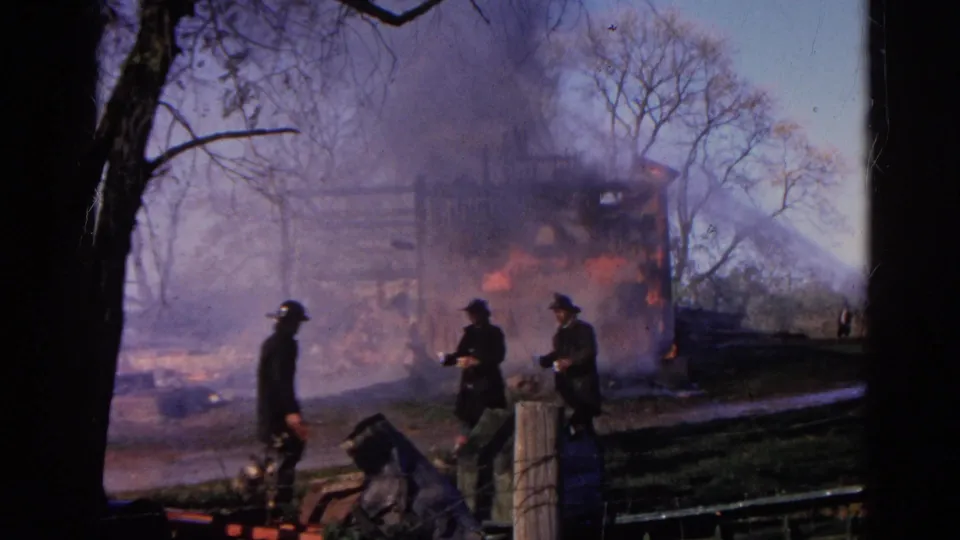 Firefighters in dark coats and helmets move across the Wauconda fire scene with flames and smoke behind them.