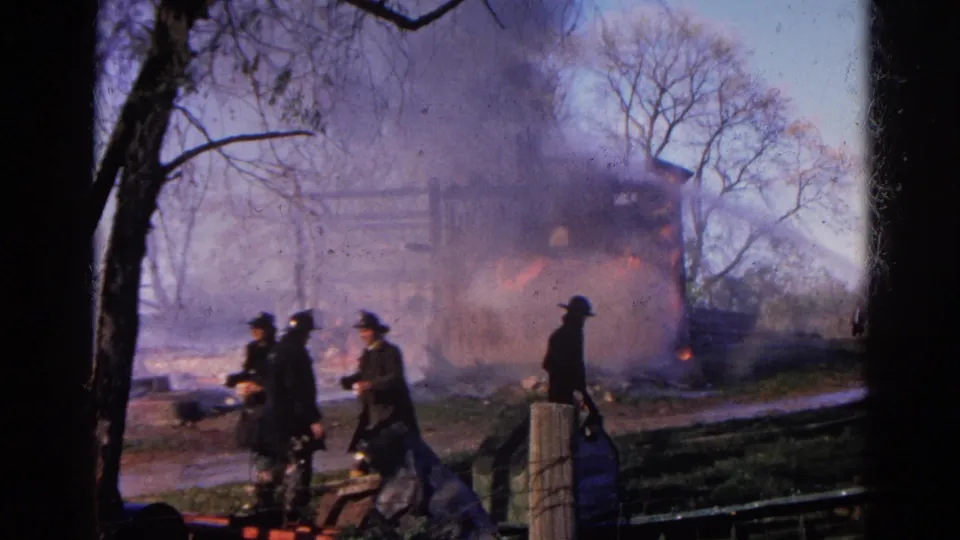 Firefighters continue working near the burning structure in Wauconda, Illinois, with smoke, flame, and bare trees visible.