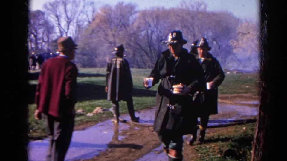 1968 Wauconda, Illinois 8mm frame showing firefighters in dark coats walking near a muddy path as smoke rises behind them.