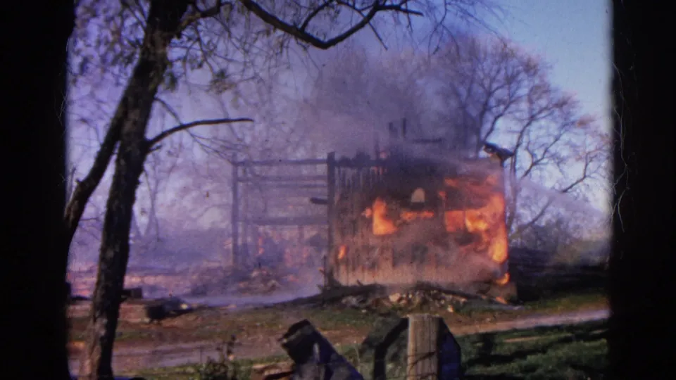 The fire continues to burn through a wooden structure in Wauconda, Illinois, framed by trees and smoky air.