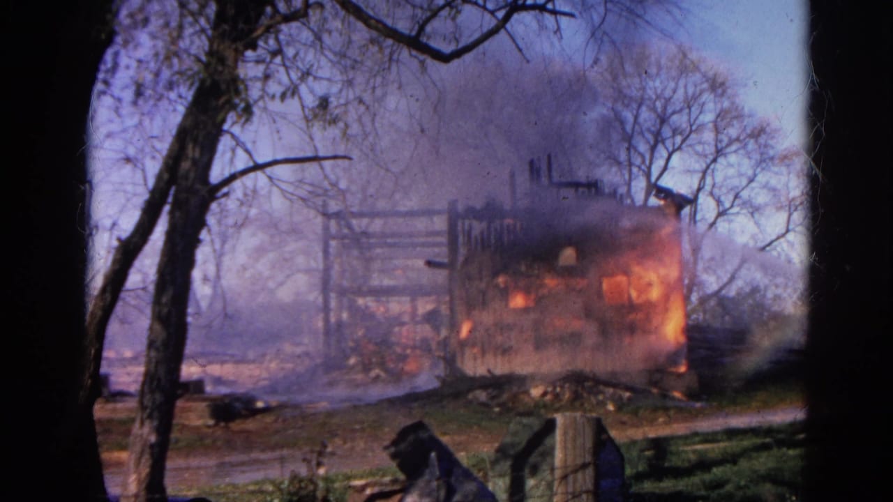 1968 Wauconda home-movie frame showing a wooden house engulfed in orange flames behind leafless trees and smoke.