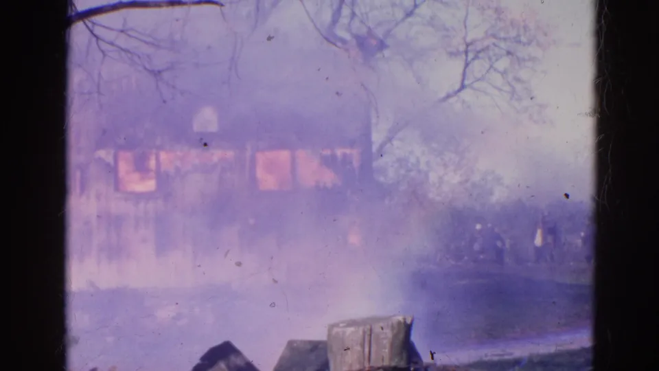 A 1968 home-movie frame shows the house fire muted by dense smoke, with fence posts and bare trees visible nearby.
