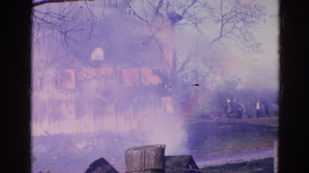 The Wauconda house fire continues through smoke, with small distant figures standing off to the right of the burning structure.