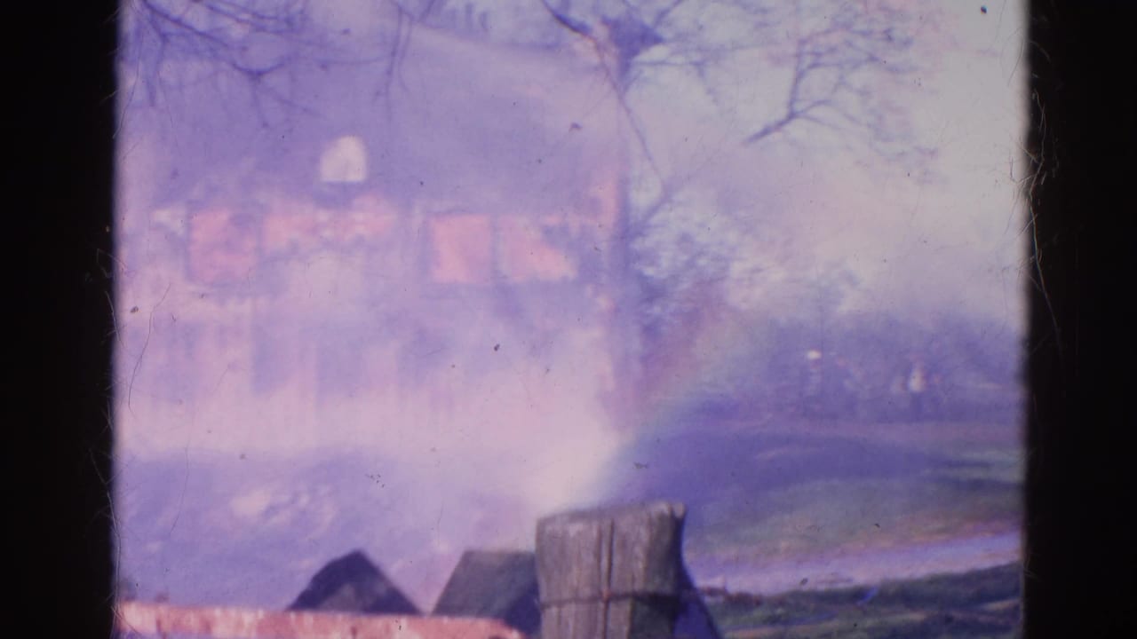 Smoke and water spray obscure a burning house beyond a wooden fence.
