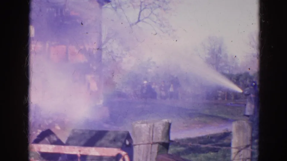 1968 Wauconda, Illinois 8mm frame with a strong water stream crossing smoke toward a house fire, with distant figures near the scene.