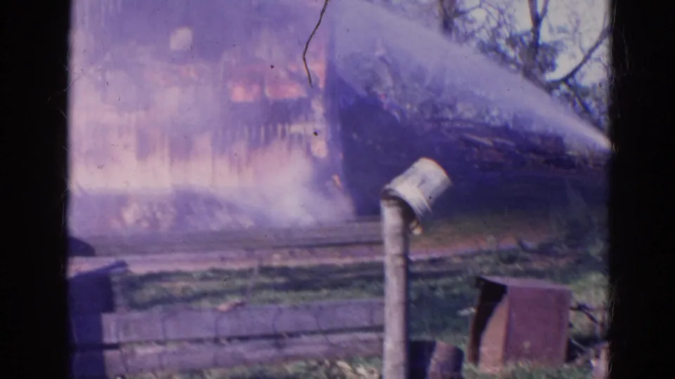 1968 Wauconda, Illinois home-movie frame showing a hose stream aimed at a smoke-covered house fire, with fence sections and a pipe in the foreground.