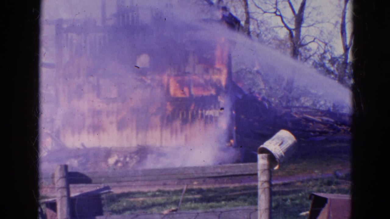Burned structure with smoke, charred wall sections, bare trees, debris, and a small post in the foreground.