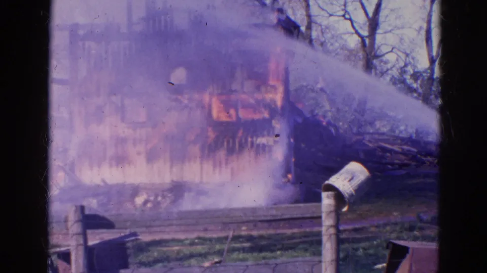 1968 Wauconda, Illinois 8mm frame showing a burned structure, smoke or haze, collapsed debris, bare trees, fencing, and a foreground post.