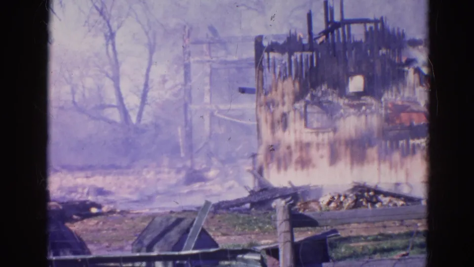 1968 Wauconda, Illinois 8mm frame showing a smoke-hazed burned wall, collapsed beams, bare trees, low fence, grass, and scattered wreckage.