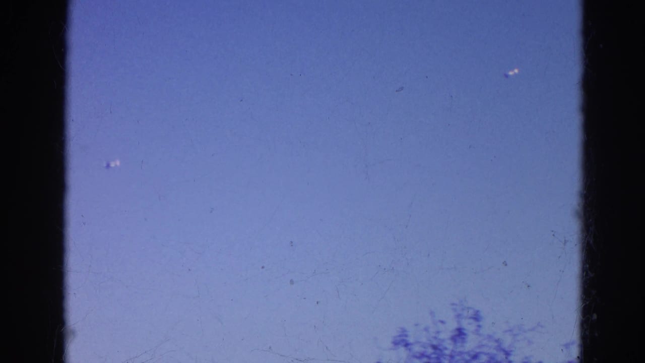 1968 8mm home-movie frame from Wauconda, Illinois, showing blue sky, dark gate edges, and tiny distant aircraft-like specks.