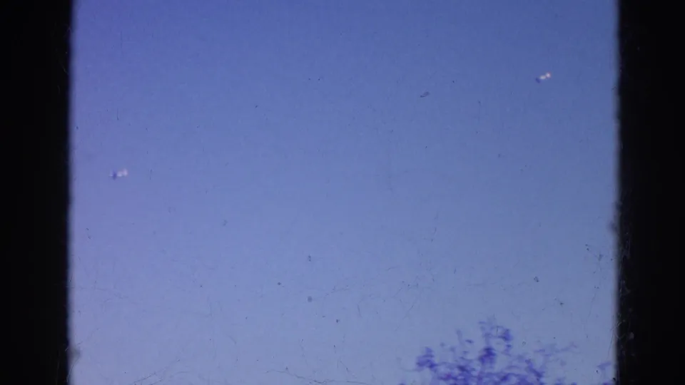 1968 8mm home-movie frame from Wauconda, Illinois, showing blue sky, dark gate edges, and tiny distant aircraft-like specks.
